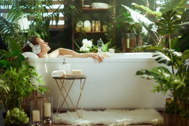 Bir kadın, yeşil ev bitkileriyle çevrili, gözleri kapalı, kulaklıkları açık, huzurlu, lüks bir banyodan zevk alır. Doğal ışık, bu sakin, spa benzeri tuvaleti doldurur. Özgüven ve sükuneti ön plana çıkarır..