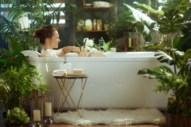 Gülümseyen bir kadın, sakin, bitki dolu bir banyoda dijital tablet kullanarak lüks bir köpük banyosunda dinleniyor. Yumuşak ışıklandırma & buhar, rahatlama ve kişisel bakım için sakin bir ev-spa atmosferi oluşturur.