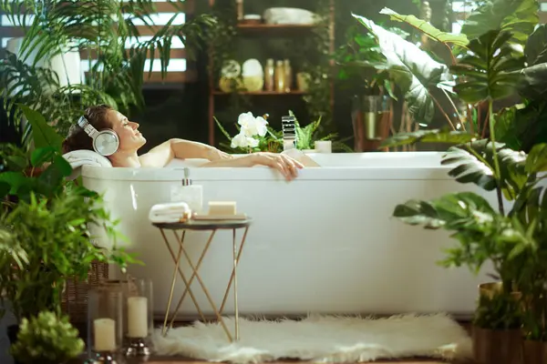 Bir kadın, yeşil ev bitkileriyle çevrili, gözleri kapalı, kulaklıkları açık, huzurlu, lüks bir banyodan zevk alır. Doğal ışık, bu sakin, spa benzeri tuvaleti doldurur. Özgüven ve sükuneti ön plana çıkarır..