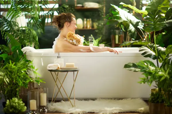 Olgun bir kadın, omzunu doğal bir banyo süngeriyle beyaz bir küvette yıkıyor. Yumuşak ışıklandırılmış banyo, yeşil bitkiler ve aksesuarlarla, barışçıl, doğal bir sağIık inzivası yaratır..