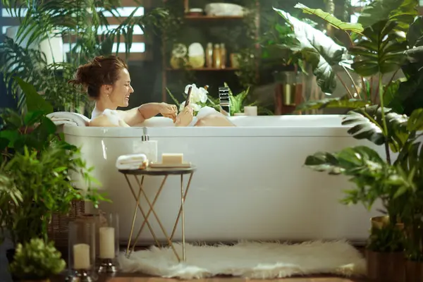 Gülümseyen bir kadın, sakin, bitki dolu bir banyoda dijital tablet kullanarak lüks bir köpük banyosunda dinleniyor. Yumuşak ışıklandırma & buhar, rahatlama ve kişisel bakım için sakin bir ev-spa atmosferi oluşturur.