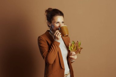 Düşünceli bir iş kadını, elinde sonbahar yaprakları olan sarı bardaktan yudumlar ve pas rengi bir ceket giyer. Sıcak, kahverengi bir arka plana doğru, sakin, samimi bir sonbahar ayarı izler..