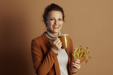 Kahverengi ceketli ve balıkçı yakalı gülümseyen bir iş kadını sarı bir kupa ve sonbahar yaprakları tutar, neşeli, memnun ve samimi bir sonbahar havasını düz kahverengi bir arka plana taşır.