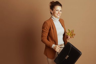 Kahverengi ceket ve bej balıkçı yaka kazak giyen kendine güvenen bir kadın elinde evrak çantası ve renkli sonbahar yapraklarıyla gülümsüyor. Sıcak kahverengi arka plana karşı duruşu profesyonel zarafeti temsil ediyor.