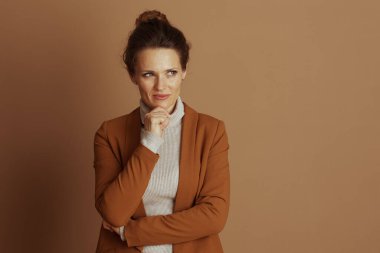 Orta yaşlı profesyonel bir kadın, eli çenesinde, düşünceli, sağa bakıyor, kafa yoruyor. Kahverengi bir spor ceket ve bej balıkçı yaka giyiyor. Kahverengi bir arka plana karşı ciddi bir tavır sergiliyor..