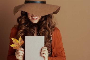 Geniş kenarlı kahverengi şapkalı ve turuncu hırkalı neşeli bir kadın elinde pembe kitap ve sarı akçaağaç yaprağı tutarak geniş bir gülümseme sergiliyor. Dalgalı kahverengi saçları bu rahat sonbahar sahnesini bej arka planda çerçeveler.