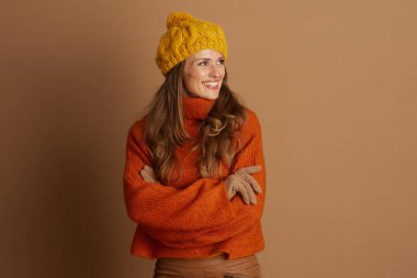 Sarı ponpon şapkalı ve turuncu balıkçı yakalı, kolları çaprazlanmış gülümseyen bir kadın yan tarafa bakıyor. Düz kahverengi bir arka planda neşeli, şık bir sonbahar ya da kış görüntüsü sunar..