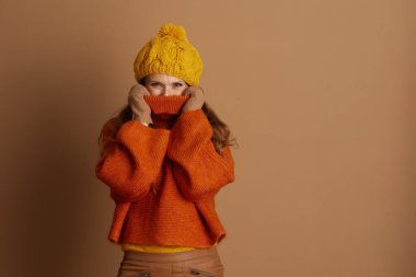 Örgü şapkalı ve büyük turuncu balıkçı yaka giyen oyuncu kadın kameraya bakarken kısmen yüzünü gizleyen eldivenli elleriyle kazak yakasını yukarı çekiyor. Bu resim sıcacık sonbahar ya da kış sıcağını yakalar