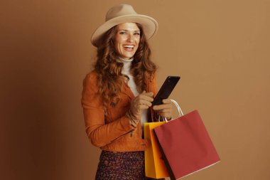 Geniş kenarlı şapka ve turuncu ceketli gülümseyen bir kadın, akıllı telefonunu ve alışveriş çantalarını tutuyor, modern sonbahar modasını ve dijital tüketiciliğini kahverengi arka planda somutlaştırıyor..