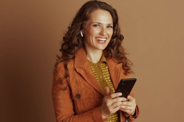 Pas rengi ceketli ve yeşil desenli bluzlu gülümseyen bir kadın kameraya neşeyle bakan bir akıllı telefon ve kablosuz kulaklık tutuyor. Neşeli, birbirine bağlı, modern bir sonbahar yaşam tarzını somutlaştırıyor..