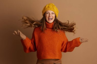 Genç bir kadın, dalgalı kahverengi saçlı, sarı bereli, turuncu balıkçı yakalı, enerjik bir şekilde gülüyor, kollarını açmış. Neşeli ifade, kahverengi arka plan, kaygısız sonbahar ruhunu yakalar..