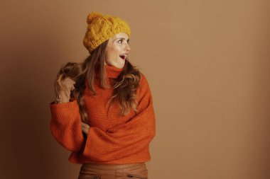 Yetişkin bir kadın, uzun dalgalı kahverengi saçlı, sarı bereli, turuncu balıkçı yaka, şaşkın ve mutlu bir ifade, saçlara dokunuyor. Orta boy, kahverengi arka plan, neşeli sonbahar enerjisi taşır..