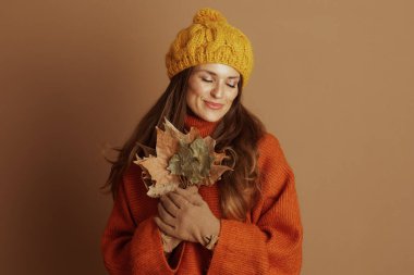 Sarı ponpon şapkalı ve turuncu balıkçı yakalı bir kadın nazikçe göğsüne dökülen yaprakları tutar gözleri kapalıdır, dingin ve rahat bir sonbahar havasını somutlaştırır..