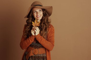 Geniş kenarlı şapkalı ve turuncu hırkalı gülümseyen bir kadın, sarı bir sonbahar yaprağını sıkıca tutmuş, sıcak kahverengi bir arka plana karşı memnuniyetle yukarıya bakıyor..