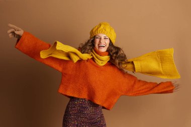 Kıvırcık kahverengi saçlı neşeli genç bir kadın parlak turuncu bir kazak, sarı örgü bir eşarp ve şapka giyer. Kolları dinamik ve kutlayıcı bir pozda uzanıyor. Sıcak kahverengi bir arka plana karşı..