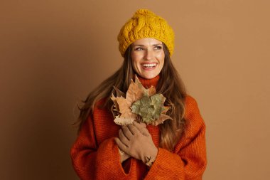 Sarı örgü şapkalı ve turuncu kazaklı neşeli bir kadın kurumuş sonbahar yapraklarını tutuyor, düzgün görünüyor. Bu rahat portre, sonbahar mevsiminin neşesini ve sıcaklığını yakalıyor. Mevsimlik temalar için mükemmel..
