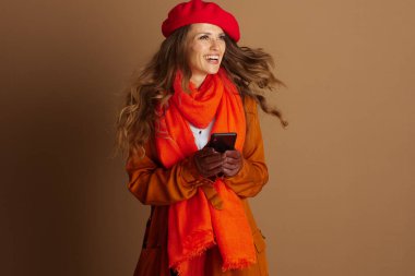 Kırmızı bereli ve turuncu eşarplı orta yaşlı bir kadın akıllı bir telefon tutarken geniş bir gülümseme sergiliyor, saçları hafif rüzgarlı. Bu stüdyo çekimi neşeli, şık bir sonbahar havasını yakalar..