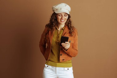 Uzun, kıvırcık kahverengi saçlı, turuncu ceketli ve bej şapkalı neşeli bir kadın akıllı telefonuna beyaz kulaklıklarla bakarken gülümsüyor..