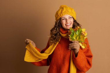 Canlı turuncu kazak, sarı şapka ve eşarp giyen neşeli bir kadın sonbahar yapraklarından oluşan renkli bir buket tutuyor, sıcak bir gülümsemeyle. Samimi giyimi ve neşeli ifadesi sonbahar eğlencesinin özünü yansıtıyor.