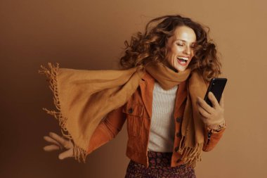 Kıvırcık kahverengi saçlı bir kadın, parlak turuncu bir ceket ve bronzlaşmış arka planda sıcak sonbahar aksesuarları giyerek akıllı telefonuna bakıp gülerken mutluluk saçar..