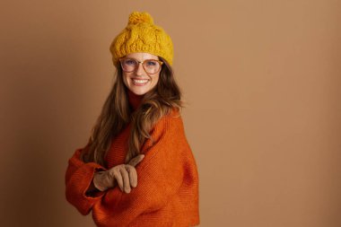 Turuncu boğazlı kazaklı, sarı bereli ve gözlüklü gülümseyen bir kadın kollarını kavuşturmuş, sıcacık sonbahar kıyafetinde sıcaklık, rahatlık ve mutluluk saçıyor..