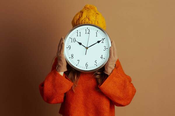 A person in a yellow beanie and orange sweater holds a large wall clock, completely obscuring their face. The image, set against a brown background, conveys themes of time or hidden identity.