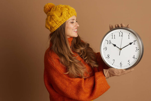 A smiling woman in a yellow beanie and orange turtleneck sweater holds a large wall clock, looking right with a cheerful expression. The scene suggests a positive embrace of seasonal time changes.