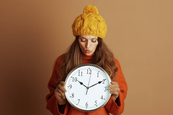 A woman in a yellow beanie and orange sweater playfully pouts, holding a large wall clock against a warm brown background. Her closed eyes suggest anticipation for seasonal changes or time adjustments