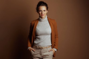 Tarafsız bir ifadeyle orta yaşlı bir iş kadını, kahverengi bir ceket ve bej pantolon giymiş, elleri ceplerinde duruyor, profesyonellik ve kendine güven saçıyor..