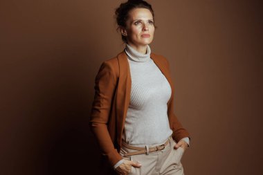 Açık turuncu ceket ve bej balıkçı yaka kazak giymiş düşünceli bir iş kadını elleri ceplerinde, yukarı bakıyor, düz kahverengi bir arka plana karşı gelecek planları tasarlıyor..