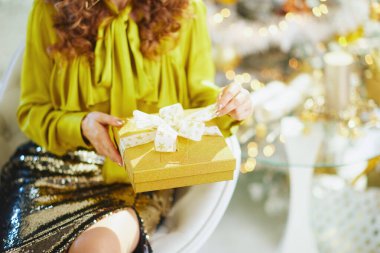 Kıvırcık kahverengi saçlı, bol sarı bluzlu ve parlak etekli zarif bir kadın elinde parlak altın bir hediye kutusuyla oturuyor. Bulanık altın bokeh ışıkları ve dekorasyonlar bayram sahnesini tamamladı.