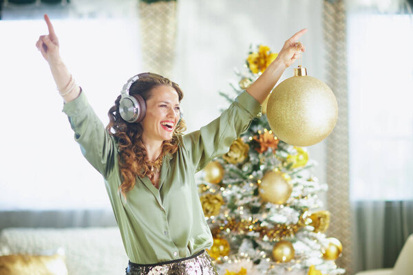 Веселая женщина с вьющимися каштановыми волосами, в наушниках и зеленой атласной блузке, весело танцует, держа в руках большое золотое рождественское украшение в праздничной, ярко освещенной комнате.