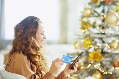Uzun saçlı gülümseyen kadın kredi kartı ve akıllı telefon tutuyor. Tatil sezonunda online alışveriş yapıyor. Parlak ışıklandırılmış, bulanık Noel ağacı neşeli ve neşeli bir ev atmosferi yaratır.
