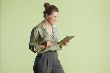 Yeşil bluzlu ve gri pantolonlu gülümseyen orta yaşlı bir kadın yeşil arka planda tabletle etkileşime geçerek modern ve profesyonel bir sahne yaratıyor..