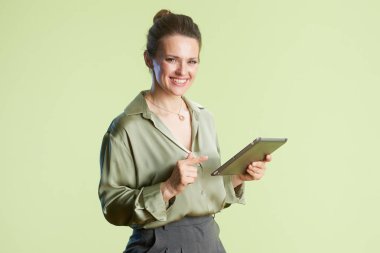 40 'larında gülümseyen bir kadın bir tablet sunar. Yeşil bluz ve gri pantolon giyiyor, açık yeşil arka planda, profesyonel, dost canlısı bir hava yaratıyor..