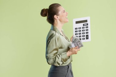 40 'lı yaşlarda elinde para ve hesap makinesi olan bir kadın gülüyor. Açık yeşil ipek bluz ve gri pantolon giyiyor, açık yeşil bir arkaplanda..