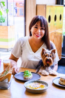 Gülümseyen genç bayan, Yorkshire teriyerinin eşlik ettiği bir hayvan dostu restoranda yemeğin tadını çıkarıyor.