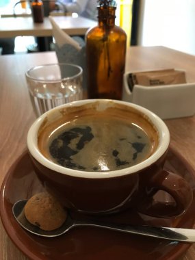 Kahverengi fincan ve çay tabağında demlenmiş espresso ile rahat ve sade bir masa üstü kahve sahnesi. Yanında bir kaşık var. Boş şişe. Ve ahşap bir masada sabah içeceği