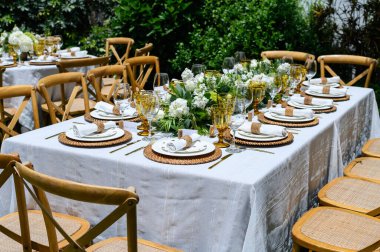 Beyaz çiçekler ve rattan detaylarla dolu bir bahçe partisi için şık bir sofra hazırlanıyor.