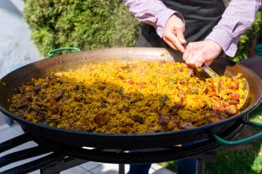 Şef, güneşli bir bahçede büyük bir paella ile metal kaşığı karıştırıyor.