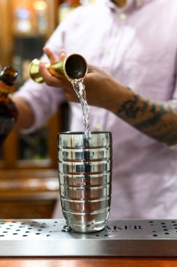 Bir barmen barda ferahlatıcı bir kokteyl hazırlıyor, profesyonel teknikler sergiliyor. İçinde paslanmaz çelik karıştırıcı, dökülen likör ve şık bir bar atmosferi var. Karışım ve içki hazırlama ile ilgili kavramlar için ideal.