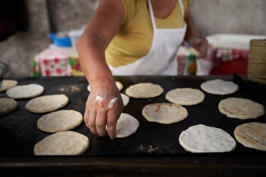 Tanınmayacak kadar yaşlı bir kadın, mütevazı mutfağındaki ızgaranın üstünde elle mısır tortillası hazırlıyor.