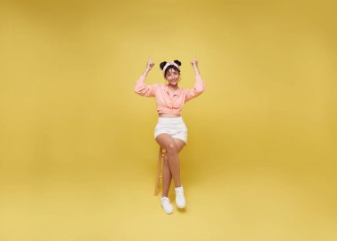 Mutlu genç Asyalı kadın gülümsüyor ve sandalyeye oturuyor. Parlak sarı arka planda izole edilmiş parmağını işaret ediyor..