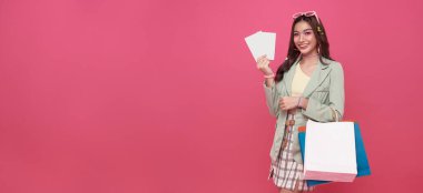 Genç gülümseyen mutlu Asyalı kadın alışverişi seviyor, alışveriş torbaları ve pembe arka planda izole edilmiş hediye kuponları taşıyor..