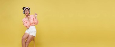 Mutlu genç Asyalı kadın gülümsüyor ve sandalyeye oturuyor. Parlak sarı arka planda izole edilmiş bir yere bakıyor ve işaret ediyor..