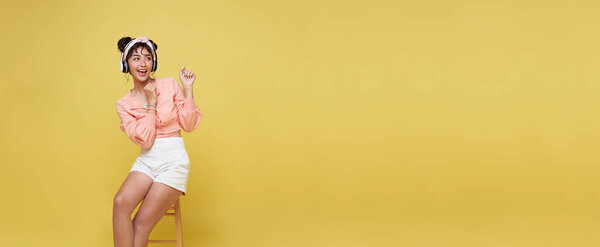 Happy young Asian teen woman smiling and sitting on chair her looking and pointing to isolated on bright yellow background.