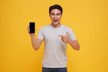 Mutlu Asyalı genç adam boş ekran akıllı telefonu gösteriyor ve işaret ediyor. Sarı arka planda izole edilmiş harika mobil teklif..