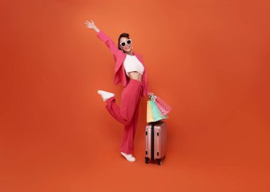 Happy young woman traveler drag luggage and shopping bag isolated on orange copy space background, Tourist girl having cheerful holiday trip concept, Full body composition