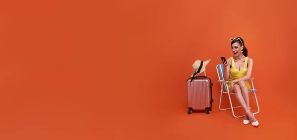 stock image happy traveler tourist woman sitting on chair with suitcase using mobile cell phone isolated on copy space orange background. Tourist girl having cheerful holiday trip concept.