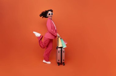 Happy young woman traveler drag luggage and shopping bag isolated on orange copy space background, Tourist girl having cheerful holiday trip concept, Full body composition
